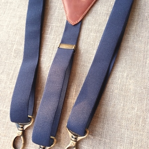 May include: Navy blue suspenders with brass-colored hardware. The suspenders feature a brown leather triangle with the initials "GS" embossed on it. The suspenders have a classic design with adjustable straps and metal clasps.