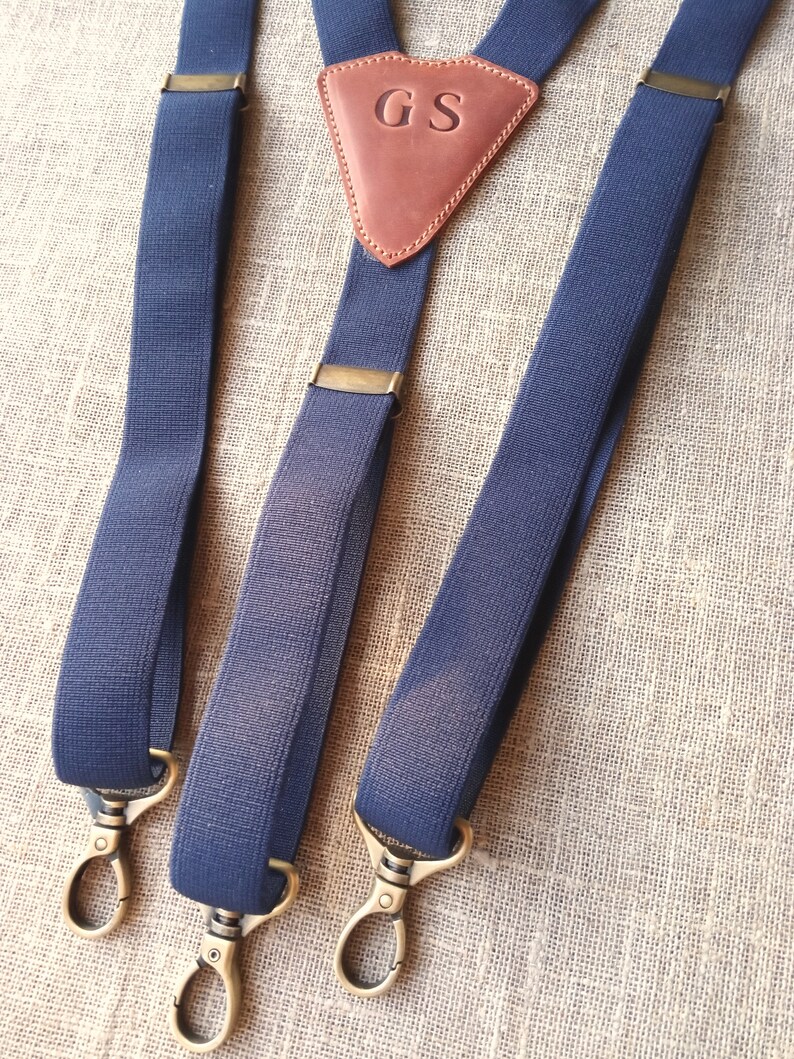 May include: Navy blue suspenders with brass-colored hardware. The suspenders feature a brown leather triangle with the initials "GS" embossed on it. The suspenders have a classic design with adjustable straps and metal clasps.