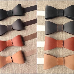May include: A collection of leather bow ties in various shades of black, brown, and tan. Each bow tie is meticulously crafted with a stitched center band and attached to an elastic strap. The bow ties are displayed on a neutral background.
