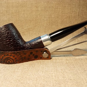 Pode incluir: Um cachimbo de madeira castanho com um bocal preto e uma banda prateada. O cachimbo está em uma caixa de couro castanho com um padrão decorativo. A caixa também inclui uma ferramenta de metal para cachimbo com um socador, um alargador e um furador.