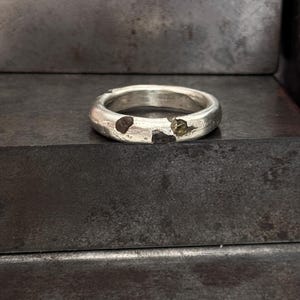 Könnte beinhalten: Ein silberner Ring mit einem einzigartigen, strukturierten Design. Der Ring hat zwei kleine, eingebettete Steine und einen absichtlich gebrochenen Abschnitt, was seine besondere Ästhetik unterstreicht. Der Ring wird auf einer dunklen, strukturierten Oberfläche präsentiert.