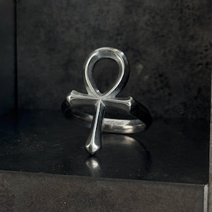 Puede incluir: Un anillo plateado con un símbolo Ankh. El anillo tiene un acabado pulido y se encuentra sobre un fondo oscuro. El Ankh, una cruz con un lazo en la parte superior, es un elemento de diseño prominente.