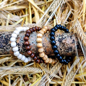 May include: Four beaded bracelets arranged in a row on a textured wooden branch. The bracelets feature spherical beads in four distinct colors: solid white, dark brown, light tan wood grain, and glossy black, each strung on a flexible cord.