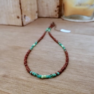 May include: A beaded necklace with a brown cord and small, round beads. The necklace features sections of turquoise and light green beads, creating a colourful accent. The necklace is displayed on a wooden surface.