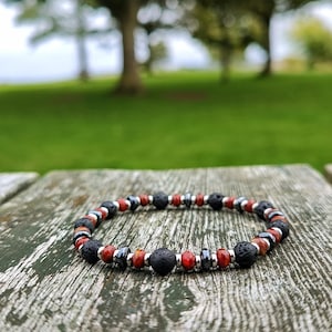 Könnte beinhalten: Ein Perlenarmband mit einem sich wiederholenden Muster aus porösen schwarzen Lavasteinperlen, glatten roten Steinperlen und kleinen metallisch silberfarbenen Zwischenperlen, die kreisförmig auf einer verwitterten Holzoberfläche angeordnet sind.