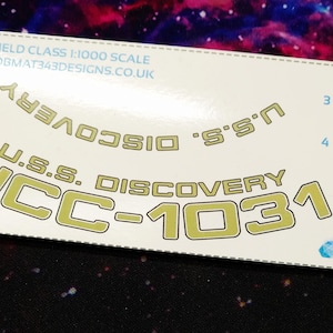 Puede incluir: Una hoja de calcomanías blanca con letras doradas que dice "U.S.S. DISCOVERY NCC-1031". El texto adicional incluye "CROSSFIELD CLASS 1:1000 SCALE" y "BOBMAT343DESIGNS". El fondo es una nebulosa colorida.