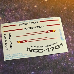 Pode incluir: Uma folha de decalque branca com marcas vermelhas e amarelas. A folha apresenta o texto "NCC-1701" várias vezes e "U.S.S. ENTERPRISE" uma vez. O texto "DECAL FILM COVERS ENTIRE SHEET - CUT DECALS ACCURATELY" está impresso no topo da folha.