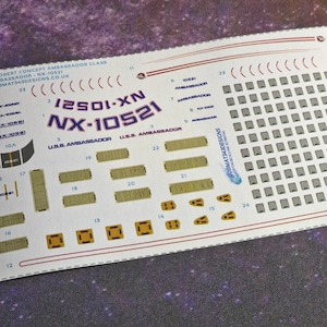 May include: A white sheet of decal paper with a variety of decals for a model starship. The decals include the text "12501-XN NX-10521", "U.S.S. AMBASSADOR", and "NX-10521".
