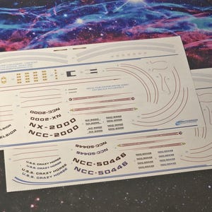 May include: Two white sheets of paper with black text and red lines. The text includes the names of starships from the Star Trek franchise, including the U.S.S. Excelsior, NX-2000, NCC-2000, and U.S.S. Crazy Horse. The sheets are designed to be used as decals for model building.