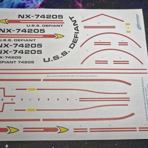 Puede incluir: Una hoja de calcomanías blanca con marcas rojas y amarillas para un modelo de nave espacial. La hoja incluye el texto "NX-74205" y "U.S.S. DEFIANT" repetido varias veces. La hoja también incluye instrucciones para "Cortar las calcomanías con precisión".