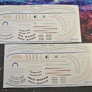 May include: Two sheets of white paper with black and blue lines and text. The top sheet has the text "U.S.S. EXCELSIOR NX-2000 NCC-2000" and the bottom sheet has the text "U.S.S. CRAZY HORSE NCC-50446".