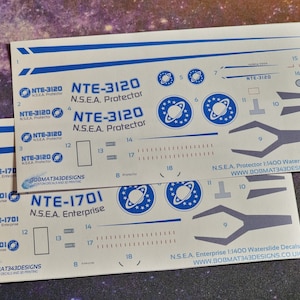 May include: Waterslide decals for a 1:1400 scale model of the N.S.E.A. Protector and N.S.E.A. Enterprise spaceships. The decals feature the ship names, registration numbers, and logos.