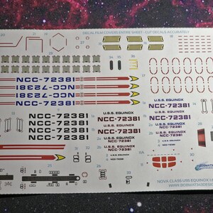 May include: A decal sheet for a model of the USS Equinox starship from Star Trek. The sheet includes the ship's registration number, NCC-72381, and other markings. The sheet is printed on white paper with red, black, and yellow accents.