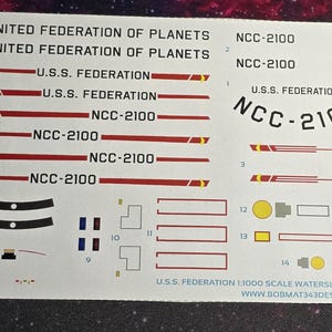 May include: A sheet of waterslide decals for model building, featuring the text "United Federation of Planets" and "U.S.S. Federation" along with the registration number "NCC-2100". The decals are printed on a white background with red, yellow, and black accents.