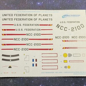 May include: A sheet of waterslide decals for a 1:1400 scale model of the USS Federation starship, NCC-2100. The decals feature the ship's name, registration number, and various markings in red, yellow, and black.