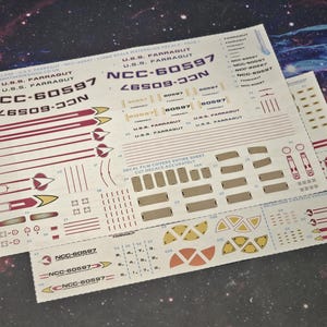 May include: Two sheets of waterslide decals for the U.S.S. Farragut NCC-60597, with markings and details. The decals are printed in red, yellow, and white on a light beige background. The text "NCC-60597" is prominently displayed.