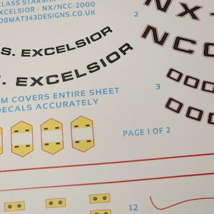 May include: A sheet of white decals with black text and red accents. The text includes "U.S.S. EXCELSIOR", "NX/NCC-2000", and "PAGE 1 OF 2". The decals feature ship names and numbers, with a few yellow and white geometric shapes.