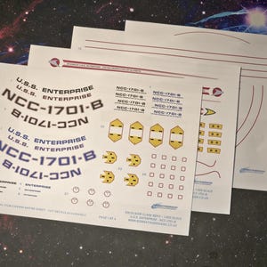 May include: Decal sheets for the U.S.S. Enterprise NCC-1701-B, with text and graphics. The sheets contain various markings, logos, and ship details. Text includes "U.S.S. Enterprise" and "NCC-1701-B".