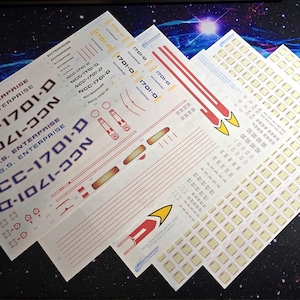 May include: A collection of decal sheets for the U.S.S. Enterprise NCC-1701-D, with text and graphics. The decals feature the ship's name, registry number, and various markings in white, red, and gold. The sheets are arranged on a dark surface with a galaxy design.