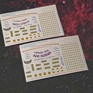 May include: Two decal sheets with text and graphics, featuring the text "NCC-1701-C" and "NX-10521". The sheets have a grid of small squares and various rectangular and square shapes. The background is a dark, speckled surface.