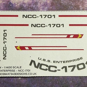 May include: A white decal sheet with red and gold stripes and the text "NCC-1701" and "U.S.S. ENTERPRISE NCC-1701". The decal sheet is for a 1:1400 scale model of the Starship Enterprise.