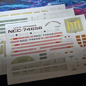 May include: Three sheets of decals for a model of the U.S.S. Voyager starship. The decals are white with black, red, and yellow details, including the ship's name and registry number NCC-74656.