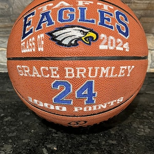 May include: A brown basketball with the text "Taft Eagles Class of 2024" and "Grace Brumley 24 1000 Points" in blue and white lettering.