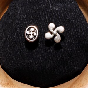 May include: Two silver charms, one with a circular design and the other with a four-pointed star design, are displayed on a black velvet cushion inside a wooden bowl.