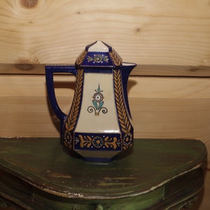 Puede incluir: Una jarra de cerámica azul y blanca con detalles dorados y diseños florales. La jarra tiene una forma geométrica y un pico en el lateral.