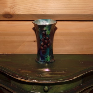 Könnte beinhalten: Eine kleine, sechseckige, schillernde Vase mit einem Traubenmuster. Die Vase ist mit einer Traube in den Farben Lila, Rot und Grün verziert. Die Vase steht auf einem grünen Tisch.