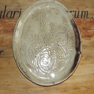 Puede incluir: Un plato de cerámica blanco con un borde marrón y un diseño en relieve de una flor estilizada. El plato está sobre una superficie de madera.