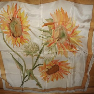 May include: A white silk scarf with a gold border features a hand-painted design of three sunflowers in shades of yellow, orange, and red.