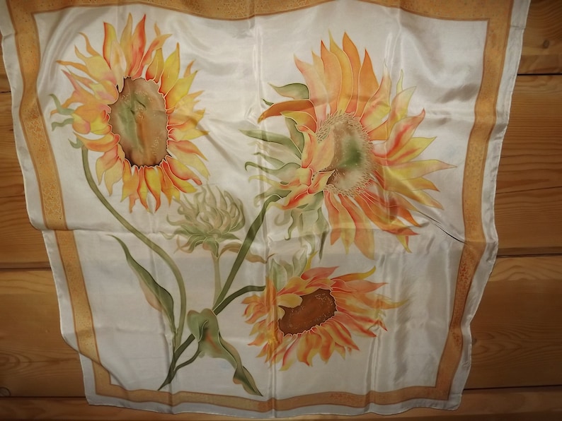 May include: A white silk scarf with a gold border features a hand-painted design of three sunflowers in shades of yellow, orange, and red.