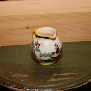 May include: A small, white ceramic creamer with a yellow rim and a hand-painted design of a woman in a red dress and a green landscape.