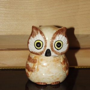 May include: A ceramic owl figurine with brown and white painted feathers, yellow eyes, and a black beak. The owl is sitting on a black surface.
