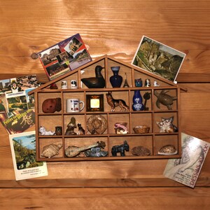 May include: A wooden display shelf shaped like a house, filled with miniature figurines and postcards. The shelf contains a variety of items, including a duck, vases, a dog, a turtle, and a guitar. Postcards with images of landscapes and buildings are also present.