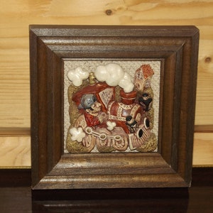 May include: A framed 3D wall art piece depicting a red and brown train with white smoke coming from the chimney. The train is on a brown and tan background with a small house in the background. The train has a figure of a person in the front.
