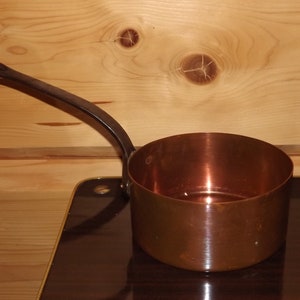 May include: A copper saucepan with a black handle. The saucepan is sitting on a dark surface.