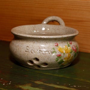 May include: A speckled beige ceramic garlic keeper with a handle. The word "Garlic" is etched on the side. Yellow and red floral designs are painted on the side. Three holes are visible near the bottom.