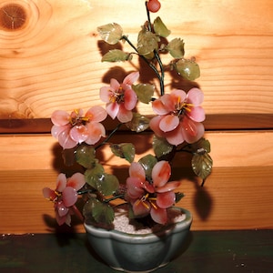 May include: A jade bonsai tree with pink flowers in a green ceramic pot. The tree is made of carved jade and the flowers are made of pink glass.