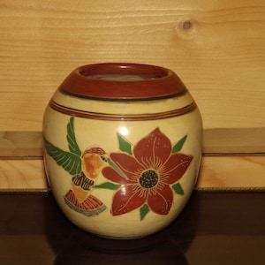 May include: A small, cream-coloured ceramic vase with a red rim and a painted design of a hummingbird and a red flower.