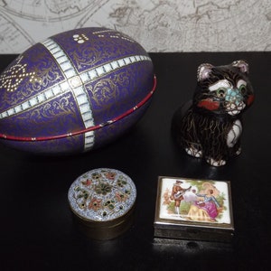 May include: Three decorative boxes: a purple egg-shaped box with gold and white details, a round brass box with a floral design, and a rectangular silver box with a painted scene of a couple.