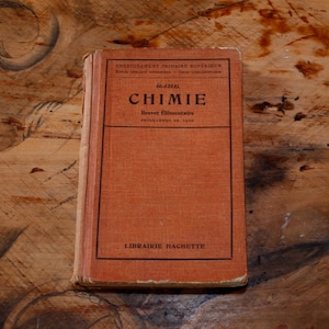 以下が含まれることがあります： 黒い文字で「CHIMIE」と書かれた、オレンジ色のヴィンテージ本。表紙には「Brevet Élémentaire」や「Librairie Hachette」など、フランス語のテキストが記載されています。木製の表面に置かれた本は、その古びた外観を示しています。