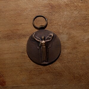 May include: A bronze medal with a ring for hanging. The medal features a raised image of a woman with wings outstretched, holding a wreath in her hands.