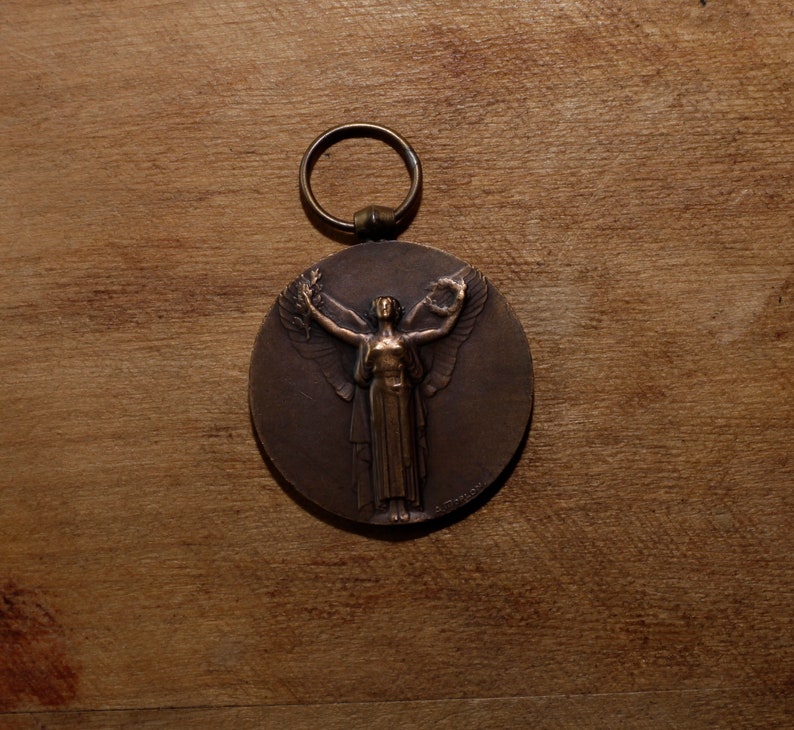 May include: A bronze medal with a ring for hanging. The medal features a raised image of a woman with wings outstretched, holding a wreath in her hands.