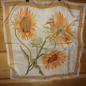 May include: A white silk scarf with a gold border and a hand-painted design of three sunflowers. The sunflowers are yellow, orange, and brown with green leaves.