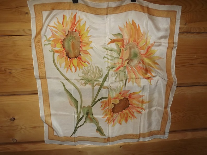 May include: A white silk scarf with a gold border and a hand-painted design of three sunflowers. The sunflowers are yellow, orange, and brown with green leaves.