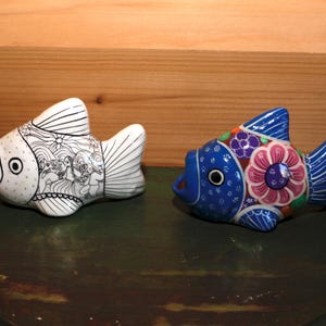 Peut inclure: Deux figurines de poissons en céramique, l'une blanche avec des détails noirs et l'autre bleue avec des motifs floraux colorés.