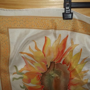May include: A white silk scarf with a hand-painted sunflower design. The sunflower is in shades of orange, yellow, and red. The scarf has a border of small orange triangles.