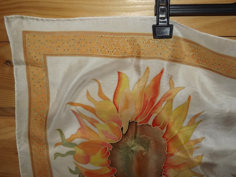 May include: A white silk scarf with a hand-painted sunflower design. The sunflower is in shades of orange, yellow, and red. The scarf has a border of small orange triangles.